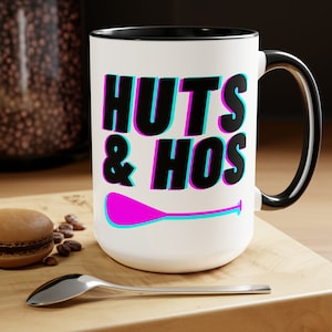 May include: White ceramic mug with a black rim and handle. The mug features a black graphic that says "HUTS & HOS" with a pink and blue paddle underneath.
