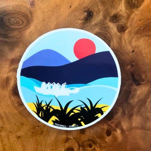 Könnte beinhalten: Ein runder Aufkleber mit einer blauen, weißen und gelben Sonnenuntergangsszene. Der Aufkleber zeigt eine Silhouette eines Bootes mit Menschen darin, Berge und eine rote Sonne. Der Text "PCG TRADING CO. JACKSONVILLE, FL" befindet sich am unteren Rand des Aufklebers.