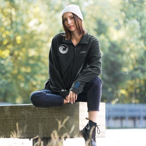 Puede incluir: Una chaqueta de forro polar verde oscuro con un logotipo de ola blanca en el pecho izquierdo. La chaqueta tiene una cremallera y un cuello alto. La modelo lleva la chaqueta con un gorro blanco y unos vaqueros azules.