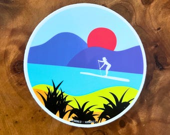 Puede incluir: Una pegatina redonda con una ilustración colorida de una persona haciendo paddle surf en un lago con montañas en el fondo y un sol rojo poniéndose. La pegatina tiene un borde blanco y un contorno negro.