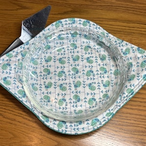 May include: A white fabric hot pad with a blue and green rooster pattern. A clear glass pie plate is sitting on top of the hot pad.
