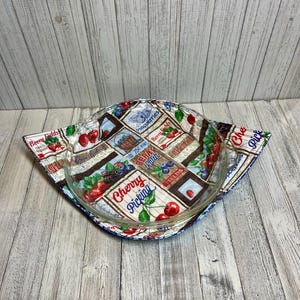 May include: A clear glass pie dish sits inside a colorful fabric bowl cozy. The fabric features a berry-themed pattern with cherries, blueberries, and the text "Cherry Picking" and "Berry Fields". The cozy is designed to protect hands from heat.
