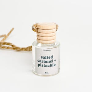 May include: A clear glass bottle with a wooden cap and a string attached. The bottle contains a car air freshener with the scent "salted caramel + pistachio". The bottle is labeled "8Twelve" and "8mL".