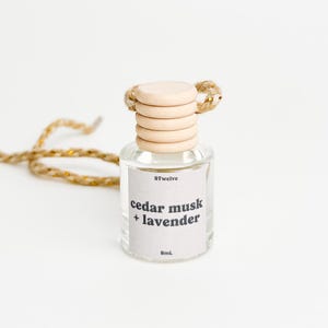 May include: A small, clear glass bottle with a wooden cap and a gold-colored rope. The label reads "cedar musk + lavender" and "8mL". The bottle is designed for aromatherapy and fragrance.