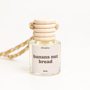 May include: A small clear glass bottle with a light-colored wooden cap and a braided cord. The label features the text 8Twelve, banana nut bread, and 8mL. The bottle contains a pale liquid and is designed for hanging as an air freshener.