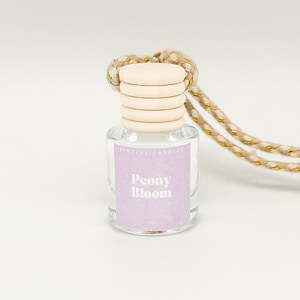 May include: A clear glass bottle with a wooden cap and a pink label that says "Peony Bloom". The bottle is hanging from a beige and gold cord.