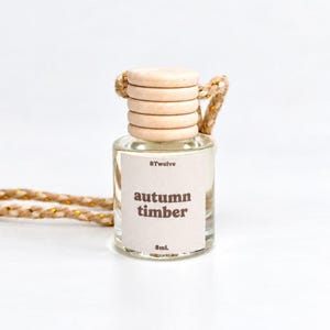 May include: A small clear glass bottle with a light wood cap and a beige label reading 8twelve autumn timber 8mL. The bottle features a braided tan cord with gold metallic threading for hanging, designed as a car air freshener diffuser.
