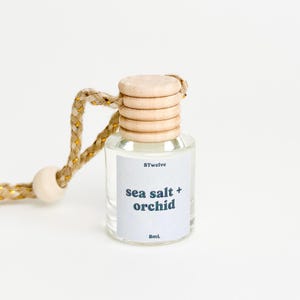 May include: A clear glass bottle with a wooden cap and a string for hanging. The bottle contains a car air freshener with the scent "sea salt + orchid". The bottle is labeled with the brand "8Twelve" and the volume "8mL".