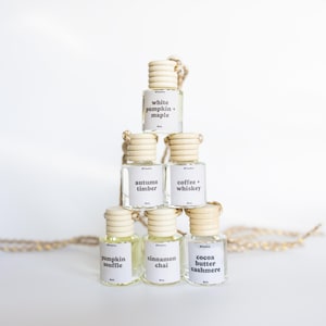 May include: A pyramid stack of six small clear glass bottles with light wood ribbed caps and hanging cords. Each bottle features a white label with black text reading White Pumpkin + Maple, Autumn Timber, Coffee + Whiskey, Pumpkin Souffle, Cinnamon Chai, and Cocoa Butter Cashmere.