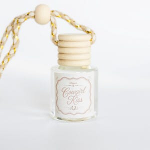 May include: A small clear glass bottle with a light wood cap and a braided cord featuring gold metallic threads. The label on the bottle has a decorative border and the text Cowgirl Kiss with a small horseshoe icon centered below the words.