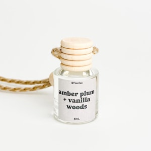 May include: A clear glass bottle with a wooden cap and a string for hanging. The bottle contains a car air freshener with the scent "amber plum + vanilla woods". The bottle is labeled "8Twelve" and "8mL".