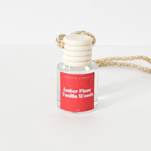 May include: A clear glass bottle with a red label that says "Amber Plum Vanilla Woods" hanging from a gold and beige string. The bottle is a car air freshener.