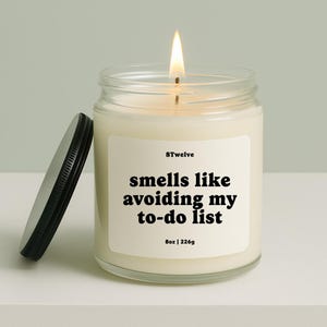 May include: A lit candle in a clear glass jar with a black lid. The label reads "smells like avoiding my to-do list" and "8oz | 226g". The candle is on a white surface.