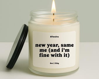 May include: A lit candle in a glass jar with a black lid. The candle has a white label with the text "new year, same me (and I'm fine with it)" and "8oz | 226g". The brand name "8Twelve" is also on the label.