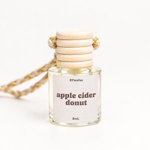 May include: A small clear glass bottle with a light wood cap and a braided rope hanger. The white label features the text 8Twelve, apple cider donut, and 8mL. The bottle contains a clear liquid and is designed for hanging in a vehicle or small space.