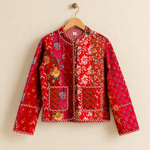 May include: A long-sleeve quilted jacket featuring a vibrant patchwork design in various shades of red, pink, and white. The jacket has a round neckline, a front button closure, two square front pockets, and white and red striped piping trim.