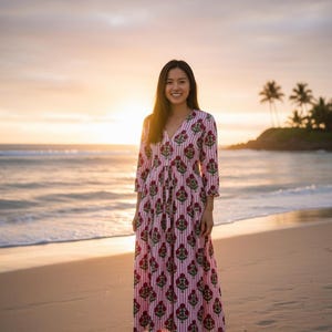 May include: A long, flowing dress with a pink and white striped pattern and floral accents. The dress has a V-neck and three-quarter sleeves. The woman is standing on a beach at sunset, with the ocean and palm trees in the background.