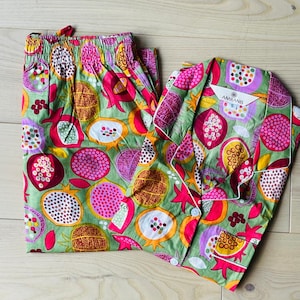 Op de afbeelding: Een tweedelige pyjamaset met een levendige granaatappeldruk in roze, groene en gele tinten. De set bevat een overhemd met knopen en witte rand en een bijpassende broek met trekkoord. De merknaam "AMRANIS" is zichtbaar.