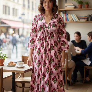 May include: A woman wearing a pink and white striped dress with a floral pattern. The dress has a V-neck and three-quarter sleeves. The background shows a cafe setting with tables, chairs, and a window.