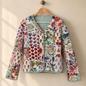 May include: A long-sleeved quilted jacket featuring a patchwork design with various floral and botanical prints in red, blue, green, and yellow. The jacket has a V-neckline, a front button closure, and green and white striped trim along the edges.