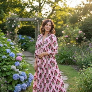 May include: A long, flowing dress with a V-neck and three-quarter sleeves. The dress features a white and pink striped pattern with a repeating floral design in a deep red color. The dress is worn outdoors in a garden setting.