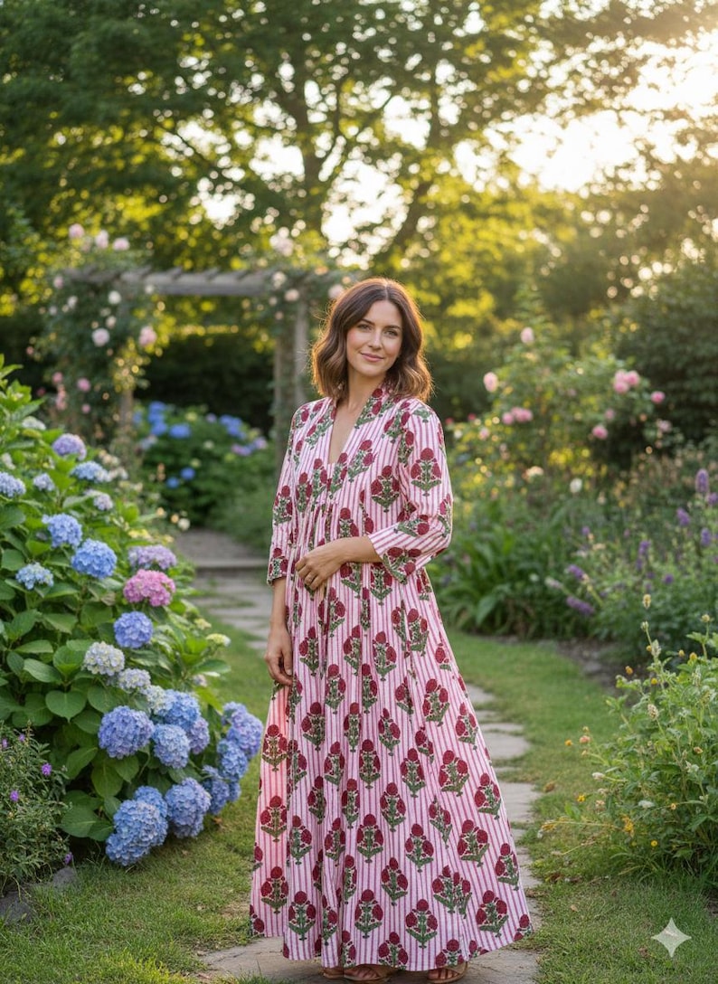 May include: A long, flowing dress with a V-neck and three-quarter sleeves. The dress features a white and pink striped pattern with a repeating floral design in a deep red color. The dress is worn outdoors in a garden setting.