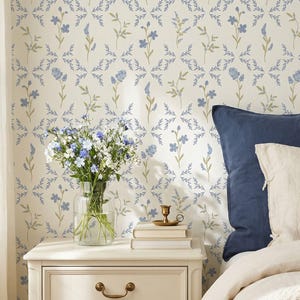 May include: A bedroom scene featuring a white nightstand with brass hardware, topped with books, a candle holder, and a vase of flowers. The bed has navy blue pillows and a cream-colored blanket. The wallpaper has a blue floral pattern.