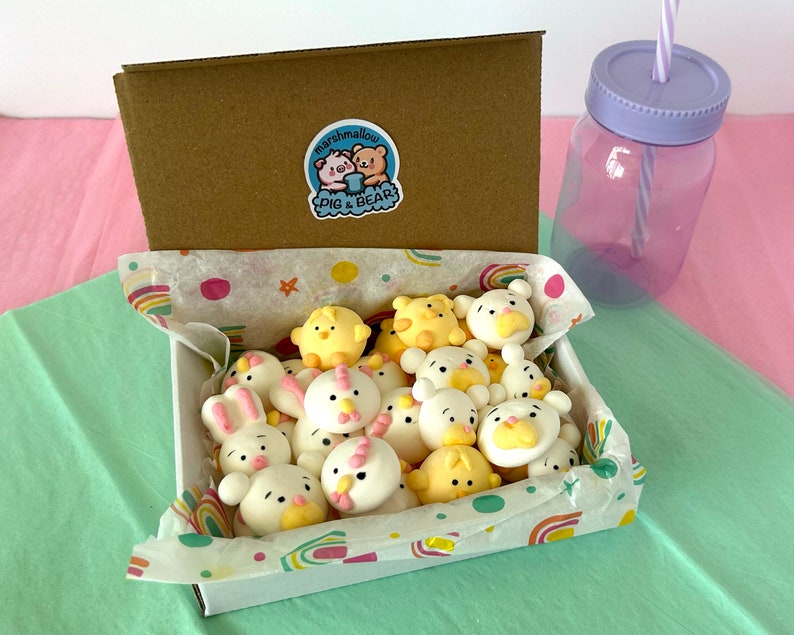 An assortment of charming marshmallow shaped like chicks, white bears, and pink-eared bunnies are presented in a box, accentuated by a colorful paper lining. A logo sticker on the box Marshmallow Pig and Bear, it delightful treat experience.