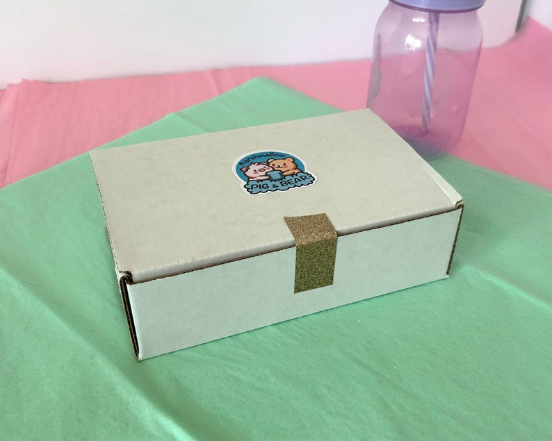 An assortment of charming marshmallow shaped like chicks, white bears, and pink-eared bunnies are presented in a box, accentuated by a colorful paper lining. A logo sticker on the box Marshmallow Pig and Bear, it delightful treat experience.