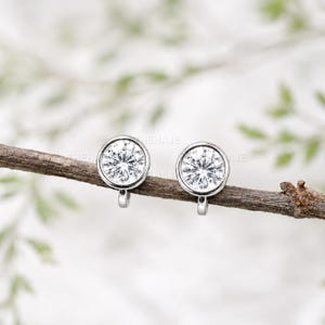 May include: A pair of silver-toned stud earrings featuring round, clear, faceted stones set in a bezel setting. The earrings are positioned side-by-side on a thin, brown, textured wooden twig, showcasing a simple and clean design.