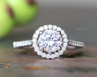 May include: A silver-toned ring featuring a large, round, clear center stone surrounded by a halo of smaller, round, clear stones. The band is also set with a row of small, clear stones along each side, creating a sparkling, multi-faceted appearance.