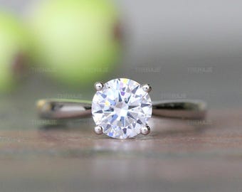 May include: A silver-toned ring featuring a single round-cut clear gemstone held in a four-prong setting. The band is smooth and polished, showcasing a reflective metallic surface. The word THEHAJE is printed multiple times in the background.