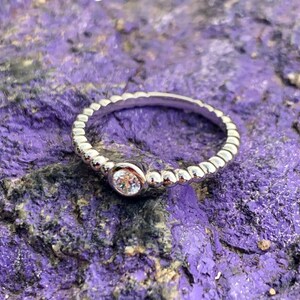 May include: A silver-toned ring featuring a beaded band design and a single, round, clear faceted stone set in a smooth bezel setting. The ring is constructed with a continuous row of small, uniform metal spheres encircling the entire band.