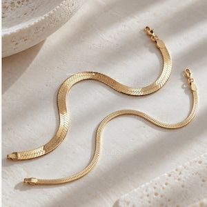 May include: Two gold-toned herringbone chain necklaces featuring a flat, woven link design. Each necklace includes a lobster claw clasp at one end and a small rectangular tab at the other. The chains are arranged in a curved, serpentine pattern.