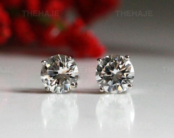 May include: A pair of round-cut stud earrings featuring clear, faceted stones set in four-prong silver-toned metal settings. The earrings are positioned side-by-side, reflecting light, with the text THEHAJE visible in the background.