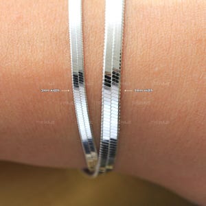 May include: Two silver snake chains, one 3mm wide and the other 4mm wide, are shown on a person's wrist. The chains are both silver and have a shiny finish.