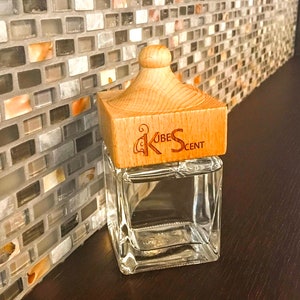 May include: A clear glass bottle with a wooden lid and the text "Kube Scent" on the lid. The bottle is sitting on a dark brown surface.