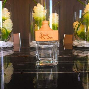 May include: A clear glass bottle with a wooden lid and the text "KOBE SCENT" on the lid. The bottle is sitting on a black surface.