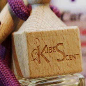 May include: A wooden car air freshener with a purple cord and a logo that says "Kube Scent" with a stylized "K".