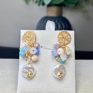 Puede incluir: Pendientes dorados con un grupo de cuentas de colores y una gran gota blanca con forma de perla. Los pendientes tienen un delicado diseño de alambre en la parte superior. Las cuentas son en tonos azules, rosas y morados.