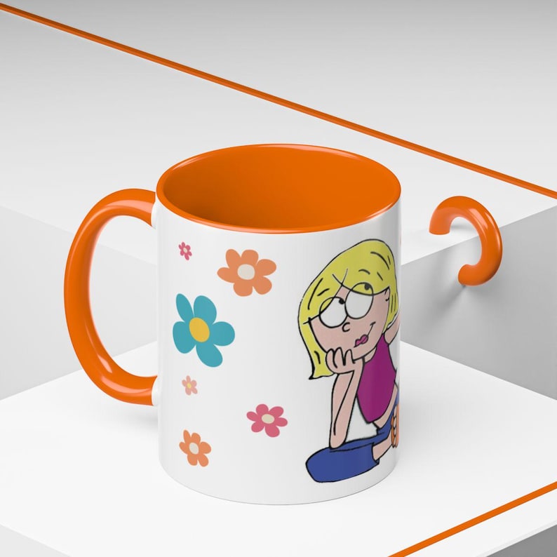 May include: A white ceramic mug with a bright orange interior and handle. The exterior features a cartoon illustration of a blonde person with a pink top and blue trousers, surrounded by small, colourful flowers in shades of blue, pink, and orange.