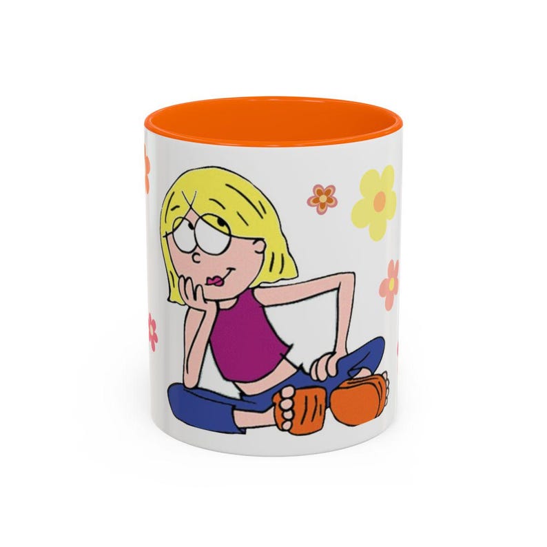 May include: A white ceramic mug with an orange rim featuring a cartoon illustration of a blonde character with a purple top and blue trousers sitting down, surrounded by small yellow, orange, and red flower graphics on the white surface.