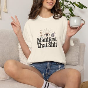 Puede incluir: Camiseta color crema con la frase "Manifest That Shit" en negro. El diseño incluye un ojo, rayos de sol y hojas. La persona lleva pantalones cortos de mezclilla y sostiene una taza.