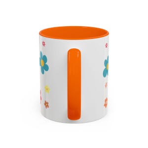 May include: A white ceramic mug featuring a bright orange handle and an orange interior. The exterior surface is decorated with a repeating pattern of small, colourful flowers in shades of blue, yellow, pink, and orange scattered across the white body.