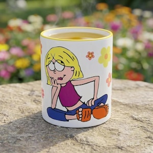 Peut inclure: Mug en céramique blanche avec un bord jaune, orné d'une illustration de dessin animé d'une fille aux cheveux blonds, un haut violet et un pantalon bleu. Le mug est décoré de motifs floraux et rempli d'un liquide clair.