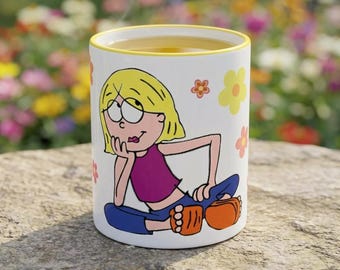 May include: A white ceramic mug with a yellow rim featuring a cartoon illustration of a blonde character with a purple top and blue pants sitting down, surrounded by small orange and yellow flower graphics on the exterior surface.