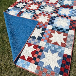 May include: A quilt featuring a repeating pattern of white stars set against a background of red, white, and blue checkered squares. The quilt has a solid blue backing with a quilted texture and a red border. Text reads: BETTY QUILT PATTERN NO. 106.