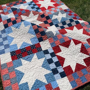 May include: A quilt featuring a repeating pattern of large white eight-pointed stars set against a background of red, white, and blue checkered and patterned fabric squares. The quilt has a visible stippled quilting texture and text reading BETTY QUILT PATTERN NO. 106 and doman design.