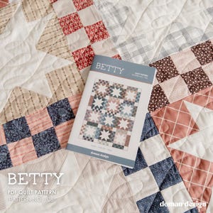 May include: A quilt pattern booklet titled BETTY, Pattern No. 106, featuring a star and checkerboard design in muted tones of red, blue, and cream. The booklet rests on a matching patchwork quilt with quilted star motifs and fabric squares.