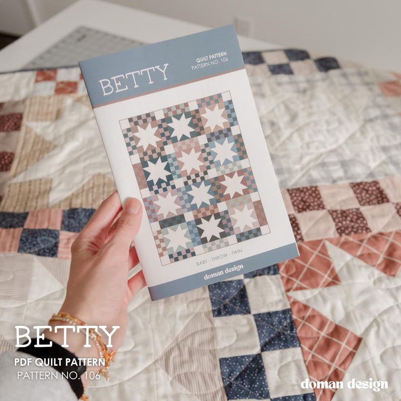 May include: A paper booklet titled BETTY Quilt Pattern No. 106 by Doman Design. The cover features a graphic illustration of a quilt with a repeating star and checkerboard block design in muted shades of blue, brown, and white, labeled Baby, Throw, Twin.